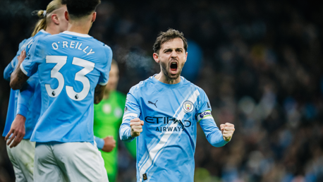 Blurred image of a Manchester City footballer celebrating a goal with teammates wearing blue shirts. One player's shirt reads 'O'Reilly 33.'