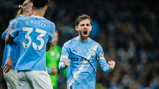 Blurred image of a Manchester City footballer celebrating a goal with teammates wearing blue shirts. One player's shirt reads 'O'Reilly 33.'