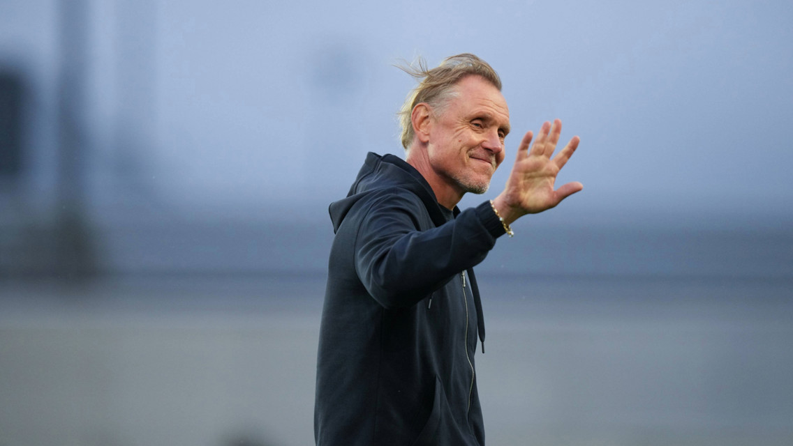 A man in a black hoodie waves with a smile.