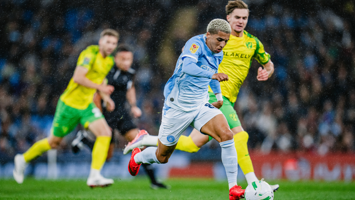 Football match scene with players wearing Manchester City and Norwich City kits. One player is in action with the ball under rain.