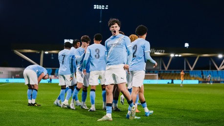 A football team in light blue jerseys with the number 3 and 2 visible, celebrating on the pitch during a night match.