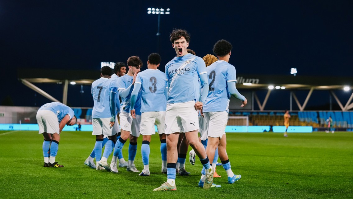 A football team in light blue jerseys with the number 3 and 2 visible, celebrating on the pitch during a night match.