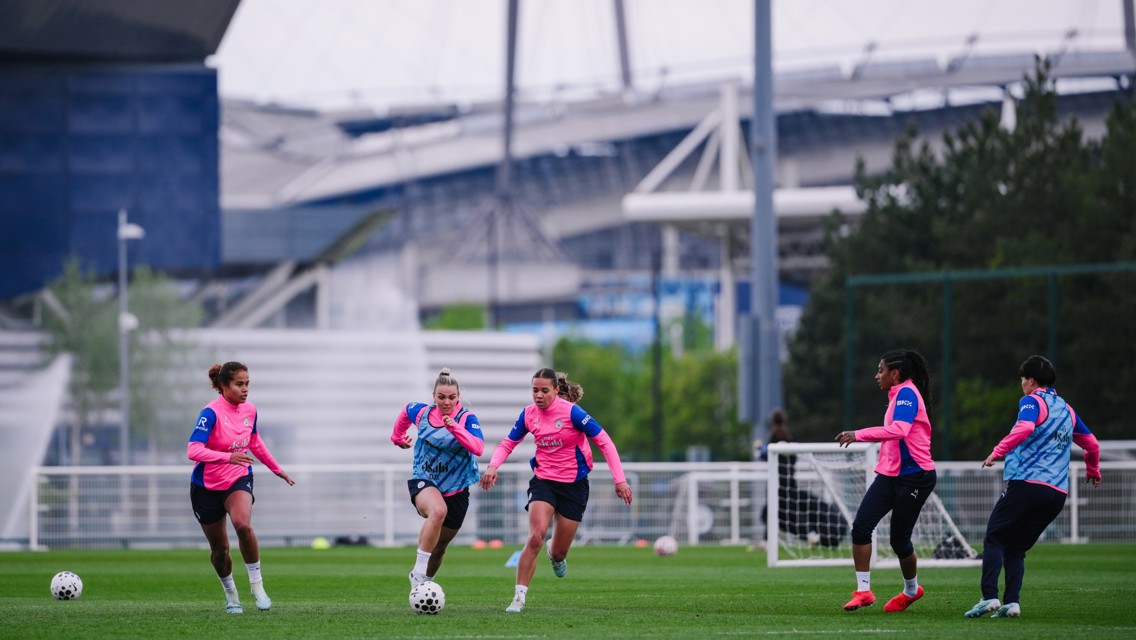 Five players in pink training tops and shorts practice football on a grass field near a stadium. There are two small goals visible and trees in the background.