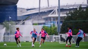 Five players in pink training tops and shorts practice football on a grass field near a stadium. There are two small goals visible and trees in the background.