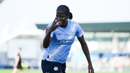A Manchester City Women player celebrates a goal on the field, wearing the team's light blue jersey and displaying a victory gesture.