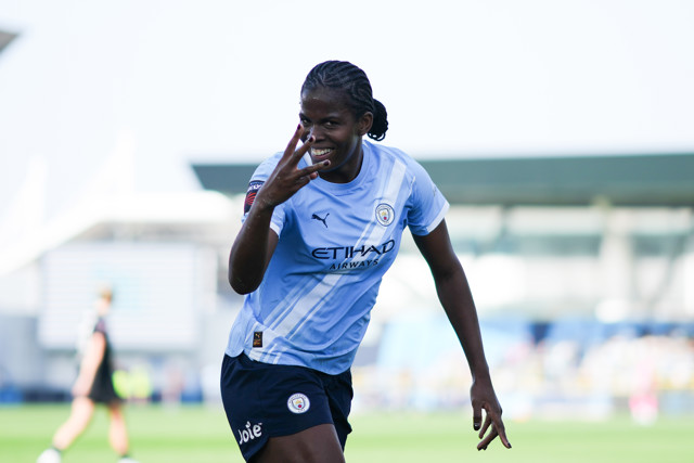 A Manchester City Women player celebrates a goal on the field, wearing the team's light blue jersey and displaying a victory gesture.