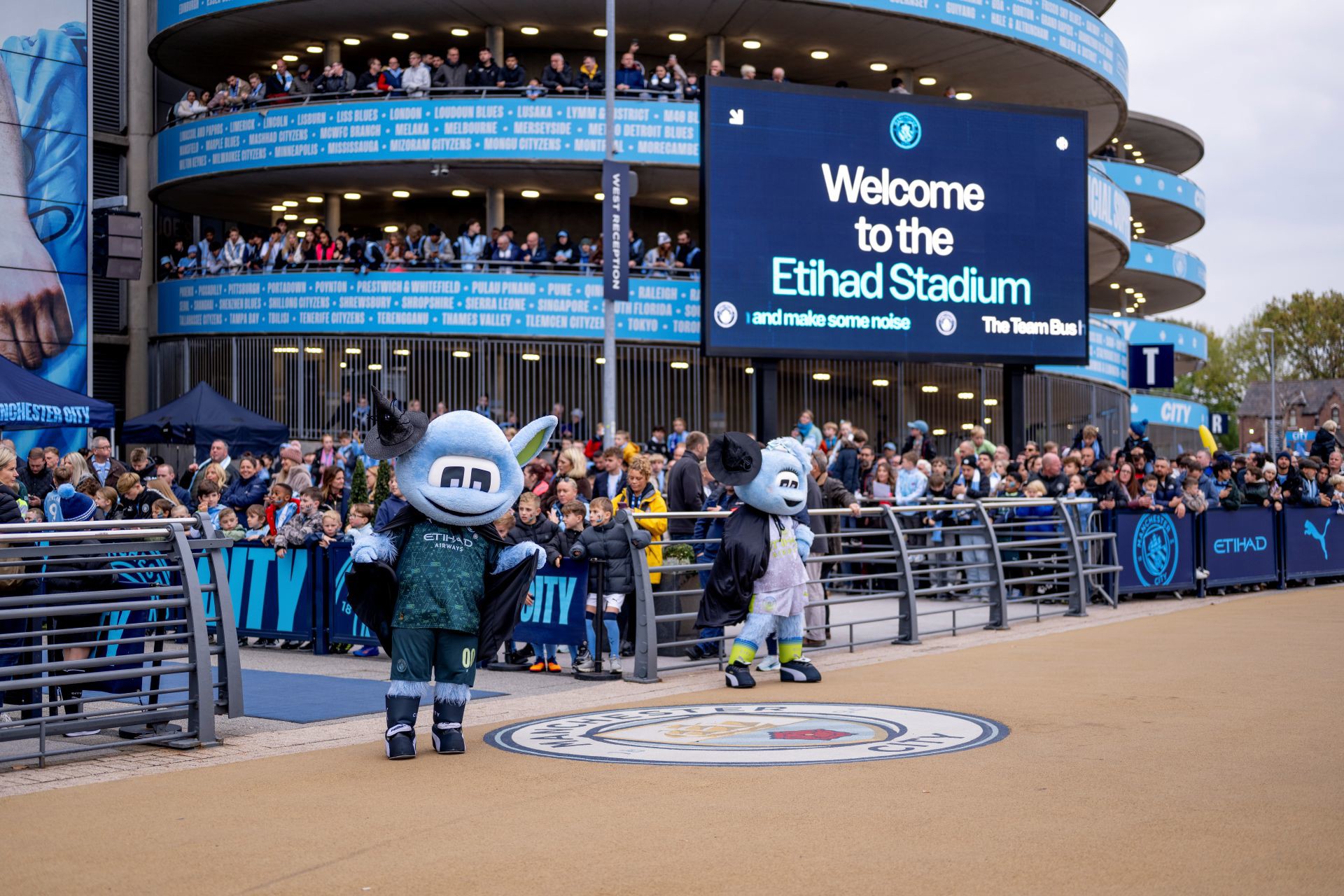 Crowd gathered at Etihad Stadium entrance with mascots and signage welcoming visitors before a Manchester City event.