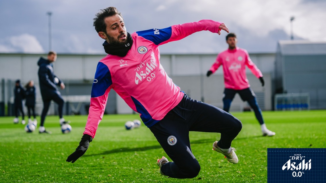 Football players in Manchester City training kits practicing on a grass field. The kits are pink with 'Asahi' branding.