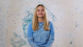 A woman with long blonde hair wearing a blue patterned shirt is sitting against a textured background.