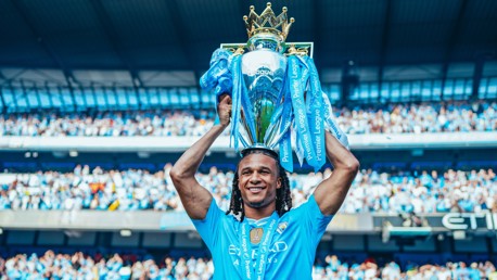 A football player in a Manchester City jersey holds the Premier League trophy aloft during a celebration at a stadium filled with cheering fans.