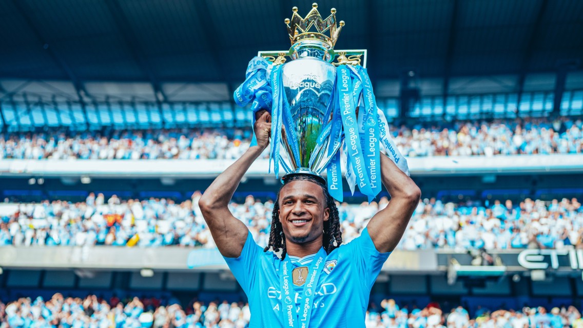 A football player in a Manchester City jersey holds the Premier League trophy aloft during a celebration at a stadium filled with cheering fans.