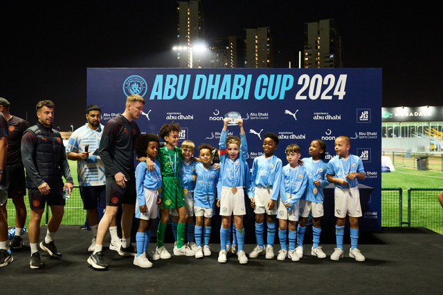 Young football players from Manchester City club standing on a stage holding a trophy during the Abu Dhabi Cup 2024 awards ceremony with coaches and staff nearby.