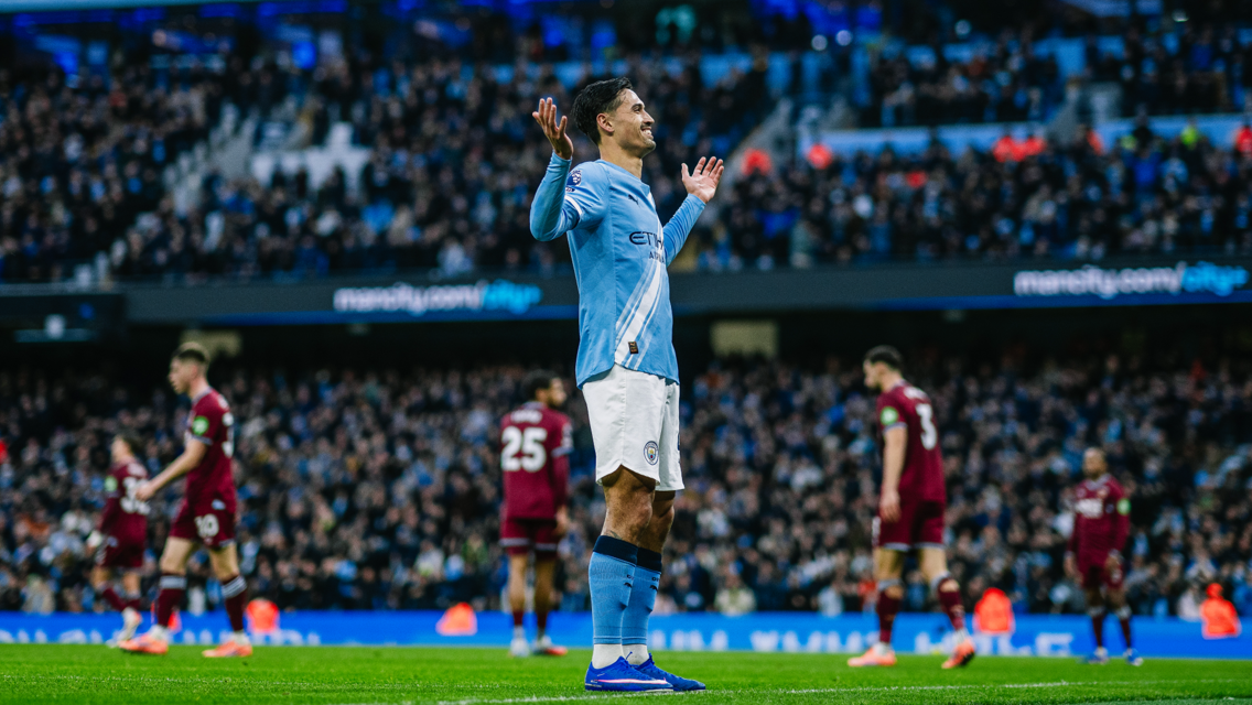 A Manchester City player in a light blue uniform gestures while on the football field during a match, with the audience visible in the background.