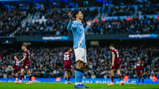 A Manchester City player in a light blue uniform gestures while on the football field during a match, with the audience visible in the background.