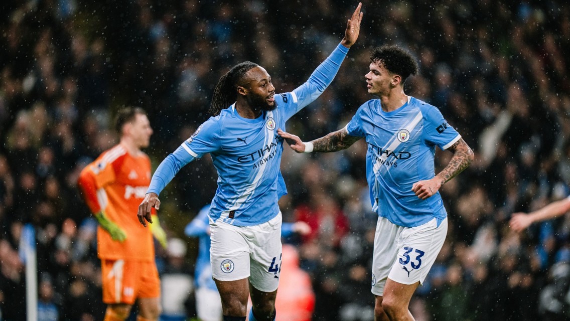 Two Manchester City players in light blue jerseys celebrate during a match in the rain, raising their hands for a high five. A goalkeeper in orange is seen in the background.
