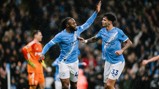 Two Manchester City players in light blue jerseys celebrate during a match in the rain, raising their hands for a high five. A goalkeeper in orange is seen in the background.