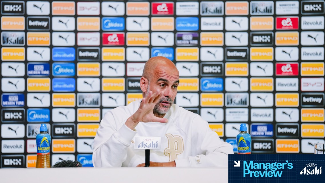 Press conference setup with blurred manager, sponsor logos, microphone labeled 'Asahi,' Lucozade bottles, and 'Manager's Preview' graphics.