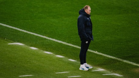 A person closely related to Manchester City stands on a football sideline wearing a dark coat and white sneakers on a grass pitch.