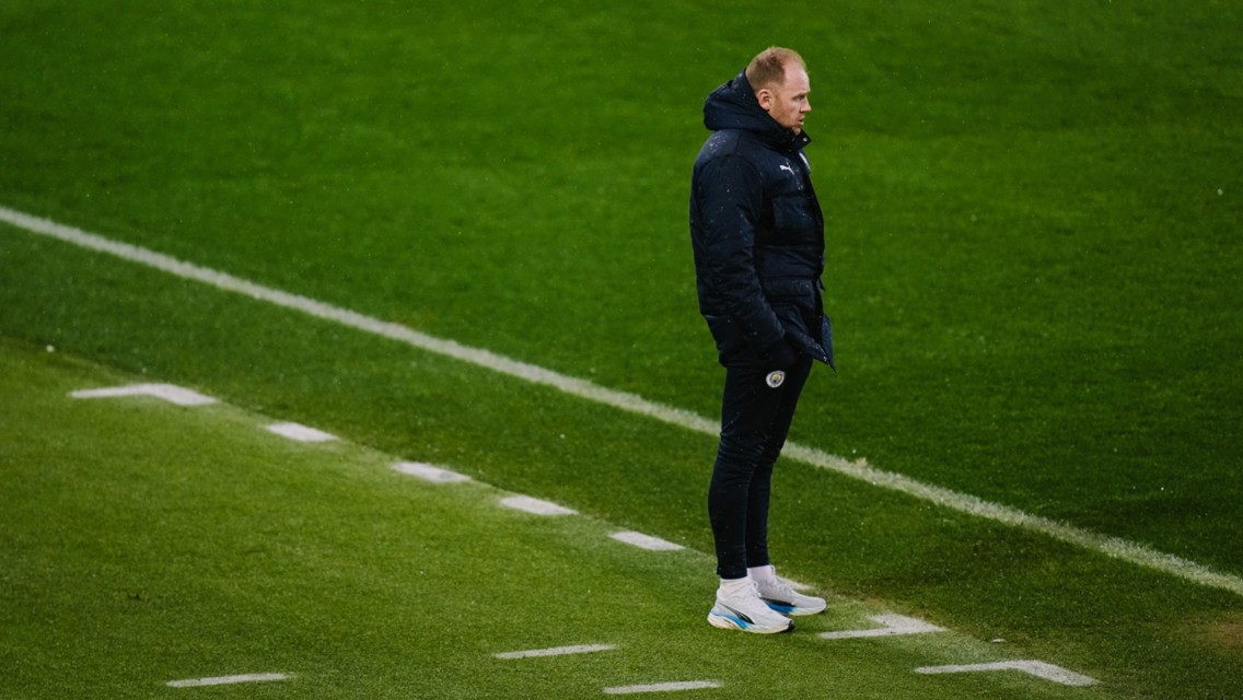 A person closely related to Manchester City stands on a football sideline wearing a dark coat and white sneakers on a grass pitch.
