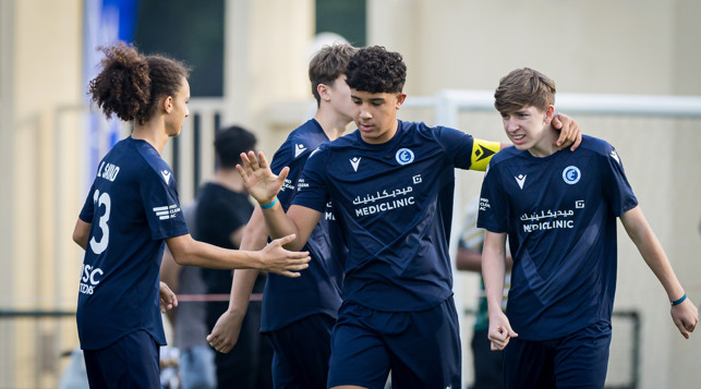 A group of young soccer players in navy blue jerseys celebrating together. The jerseys display the logo of Mediclinic and Pro Clean AC, and one player is wearing a captain's armband.