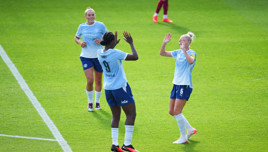 Three Manchester City Women's team players celebrate a goal on the field wearing their team jerseys.