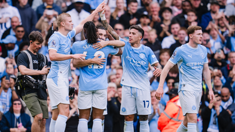 Manchester City players celebrating a goal in light blue jerseys with a crowd in the background.