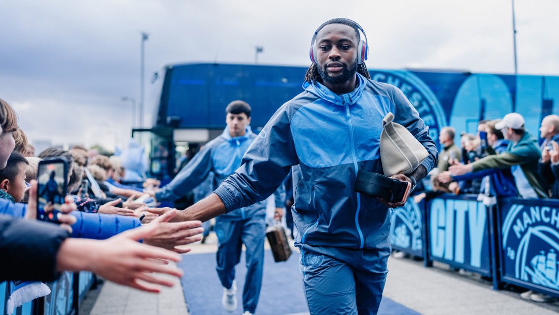 A football player wearing a blue tracksuit walks by a crowd, high-fiving fans. A team bus with Manchester City branding is in the background.
