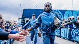 A football player wearing a blue tracksuit walks by a crowd, high-fiving fans. A team bus with Manchester City branding is in the background.