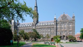 A large Gothic-style building with pointed arches and a central tower surrounded by trees and benches on a sunny day.