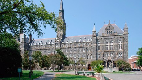 A large Gothic-style building with pointed arches and a central tower surrounded by trees and benches on a sunny day.