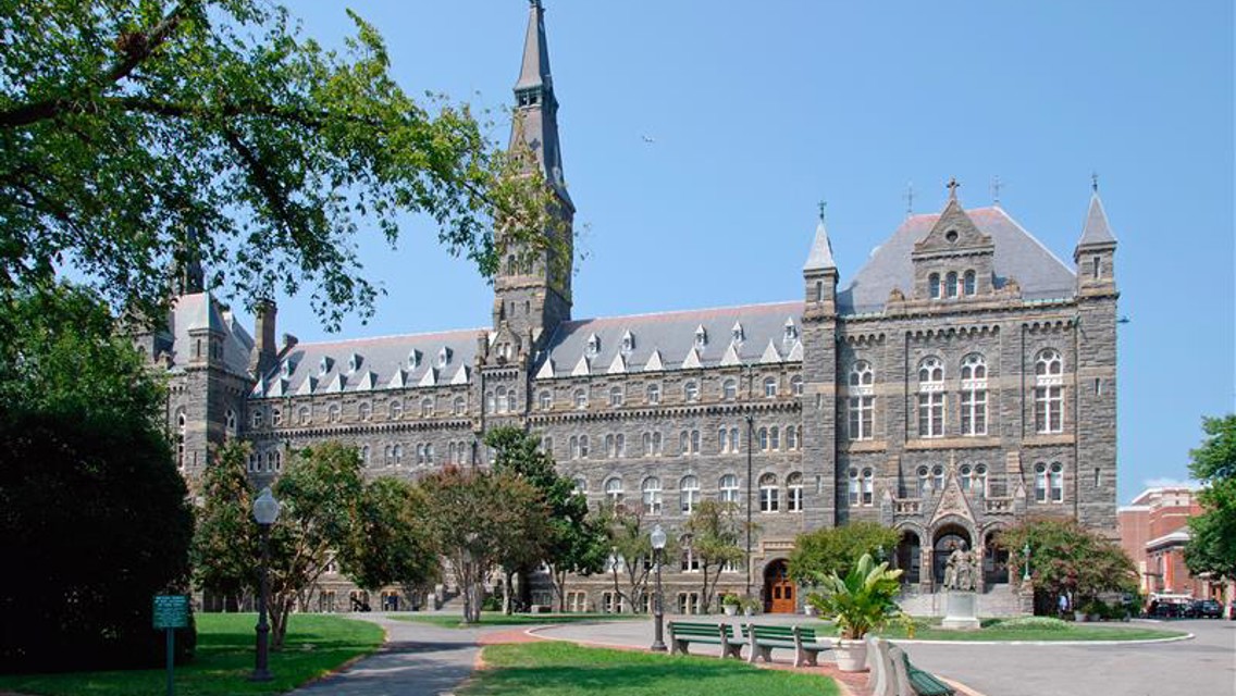 A large Gothic-style building with pointed arches and a central tower surrounded by trees and benches on a sunny day.