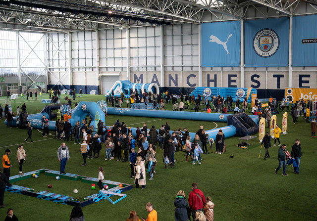 Indoor community event at Manchester City's training facility with families engaging in football activities and inflatable games. Banners display Manchester City's logo and partnership with Puma.