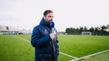 A person in a Manchester City jacket holding a microphone stands on a football field, with a blurred face. The field and goalposts are visible in the background.