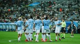 Football players in light blue jerseys, celebrating a goal on a field with a large crowd in the background.
