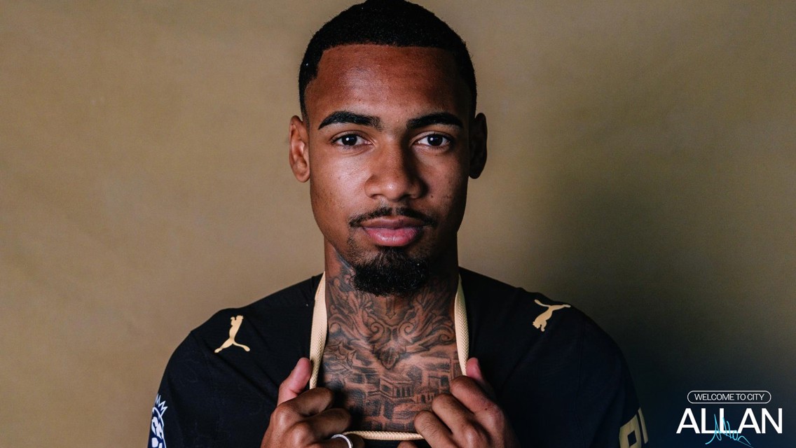 A man with tattoos poses holding the collar of a black Puma shirt.