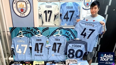 A person holds a Manchester City jersey with 'Foden' printed on it, surrounded by various Manchester City jerseys featuring 'Foden' and different numbers. A Manchester City logo is visible, and an 'Asia Tour '26' banner is present.
