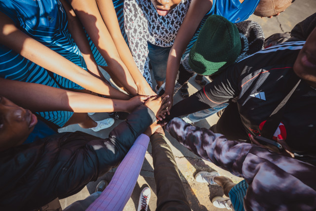 A group of people wearing casual and sportswear forming a circle and joining hands in the center, symbolizing unity and teamwork.