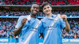 Two soccer players wearing Manchester City kits holding FA Cup medals in a stadium with crowd in background.