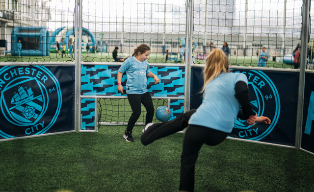 Two girls wearing Manchester City jerseys play football indoors, with one taking a shot at goal. The net and Man City logo are visible.