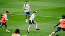 Soccer players in a training session on a grassy field, wearing white shirts and blue shorts. Some players wear orange bibs over their shirts.