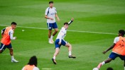 Soccer players in a training session on a grassy field, wearing white shirts and blue shorts. Some players wear orange bibs over their shirts.