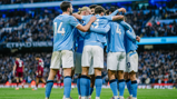 A group of Manchester City football players in a team huddle on the field, wearing sky blue and white uniforms with player numbers visible.