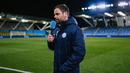 Person holding a microphone with Manchester City logo in a stadium at night.
