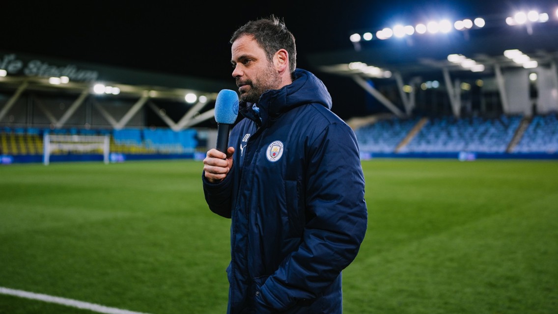 Person holding a microphone with Manchester City logo in a stadium at night.