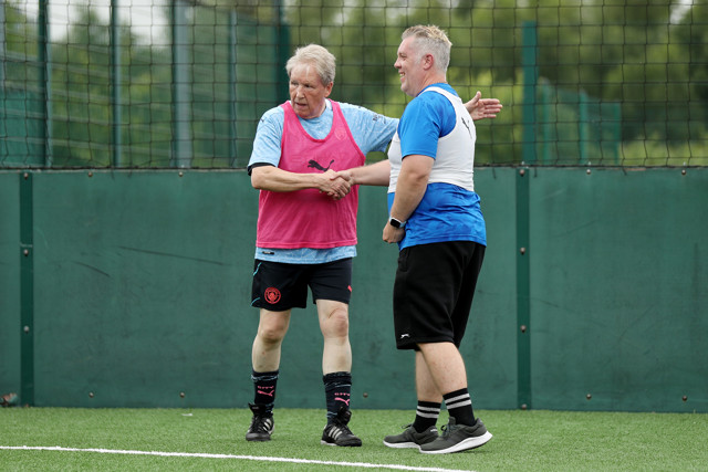 Two men shaking hands on a football field. One is wearing a pink bib and the other a white bib during a training session.