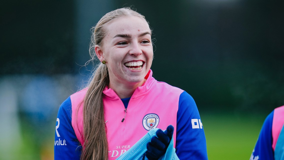 A Manchester City player wearing a pink and blue training jersey with the Manchester City logo during a practice session, holding gloves. The player's face is blurred.