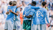 A group of Manchester City players in blue jerseys are hugging and celebrating on the field. The jerseys display names and numbers. The background is blurred with a crowd.