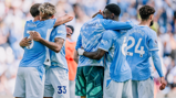 A group of Manchester City players in blue jerseys are hugging and celebrating on the field. The jerseys display names and numbers. The background is blurred with a crowd.