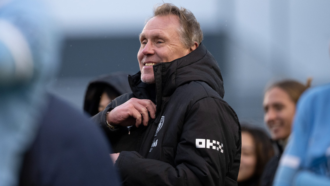 A person wearing a black coat with an OKX logo on the sleeve, possibly a Manchester City coach or staff member, standing outdoors in a group.