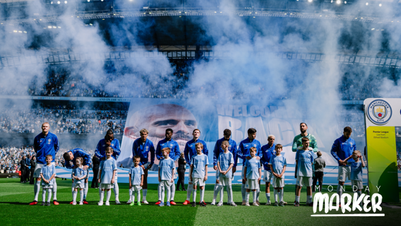 Manchester City players line up before a match at Etihad Stadium with a large banner in the background and children in Manchester City shirts.